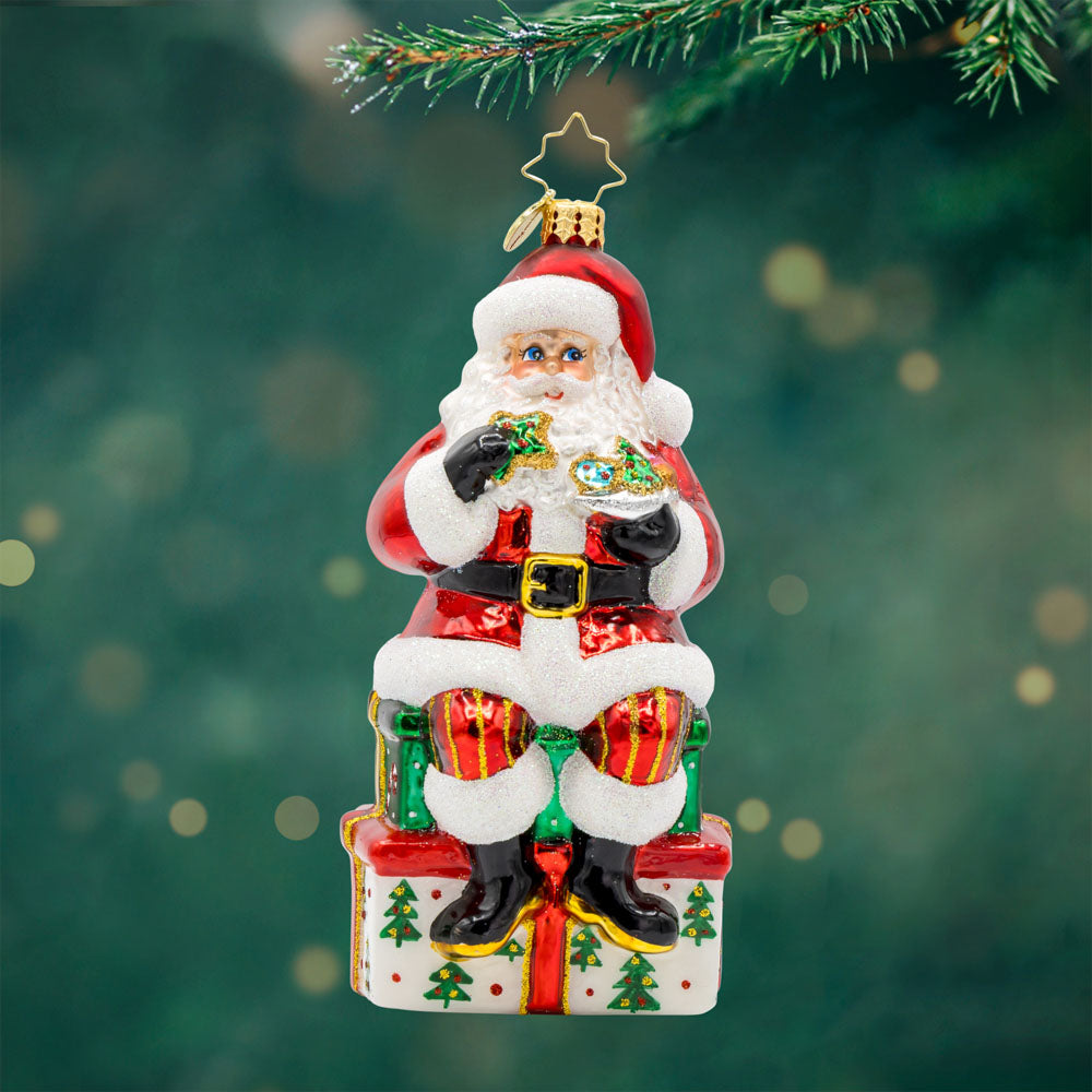 Kris Kringle's Cookie Break ornament. Santa takes a cookie break, perched atop a glittering present. View of front.