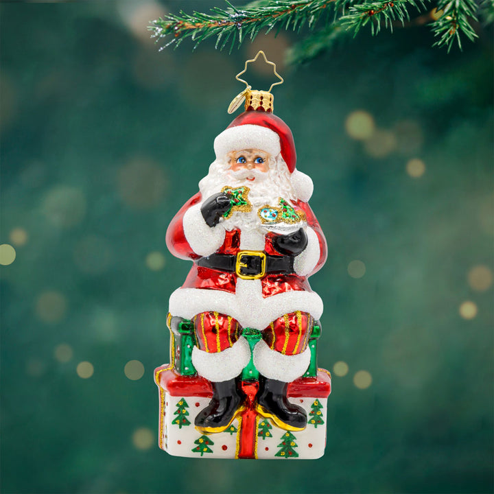 Kris Kringle's Cookie Break ornament. Santa takes a cookie break, perched atop a glittering present. View of front.