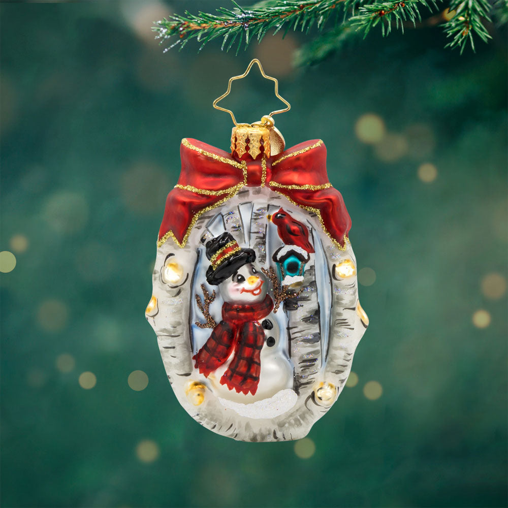 A Snowy Feathered Friend Gem ornament. A snowman and a cardinal in its birdhouse, are encircled by a wreath made of birch branches with a red bow on top. View of front. 