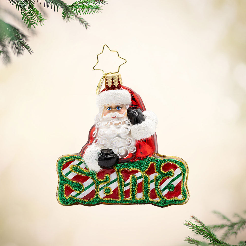 What’s in A Name Santa Gem ornament. Santa leans atop a green and red striped festive "Santa." View of front. 