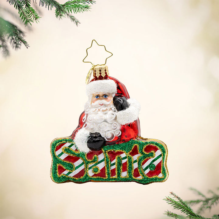 What’s in A Name Santa Gem ornament. Santa leans atop a green and red striped festive "Santa." View of front. 