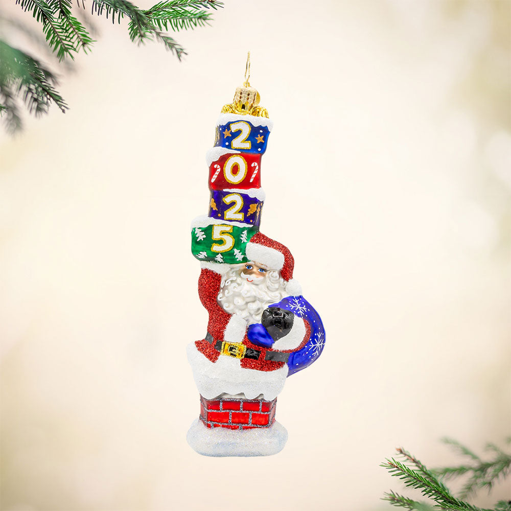 Kris Kringle's Chimney Trek 2025 ornament. Santa is coming out of a chimney, gift sack in hand and a stack of gifts dated "2025." in the other. View of front.