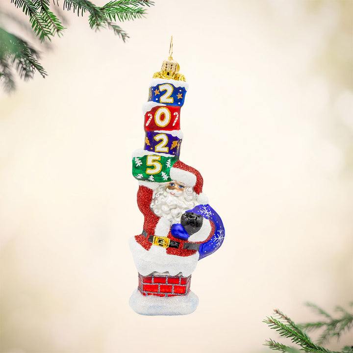 Kris Kringle's Chimney Trek 2025 ornament. Santa is coming out of a chimney, gift sack in hand and a stack of gifts dated "2025." in the other. View of front.