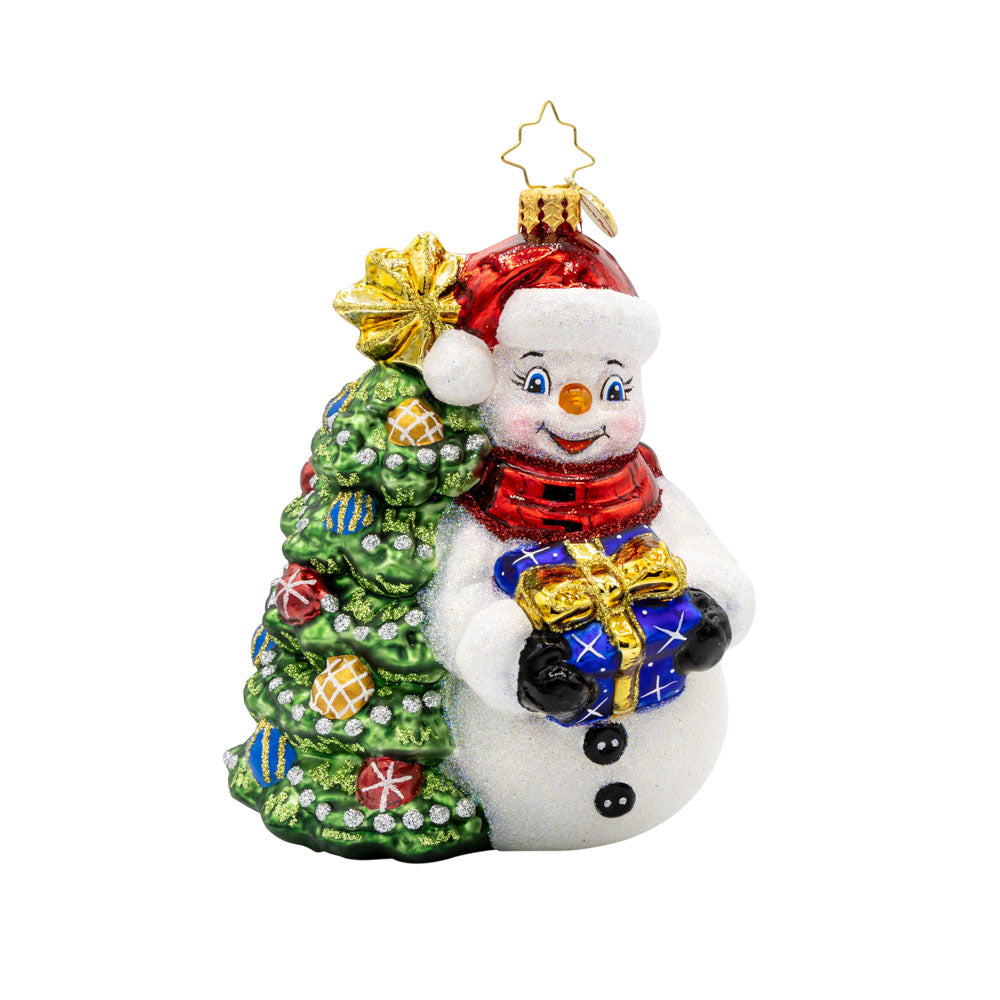 Snowy & Bright ornament. This snowman dons a red Santa hat, bearing a gift in hand. He leans against a decorated Christmas tree. View of front.