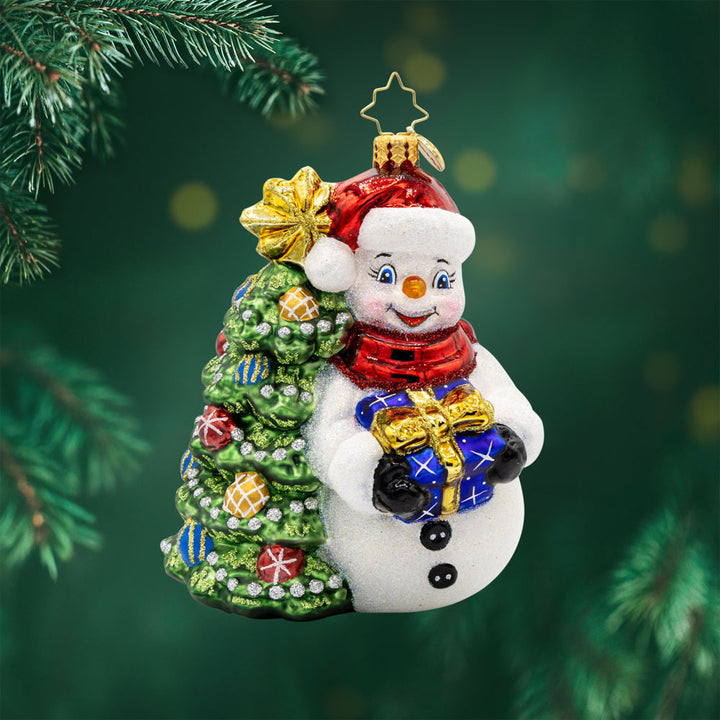Snowy & Bright ornament. This snowman dons a red Santa hat, bearing a gift in hand. He leans against a decorated Christmas tree. View of front.