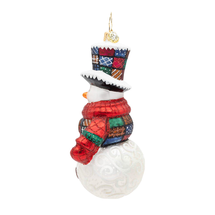 The side of a snowman Christmas ornament with a white textured body, a red glittery scarf, a checkered top hat, and a colorful patchwork-patterned vest, shown on a plain white background.