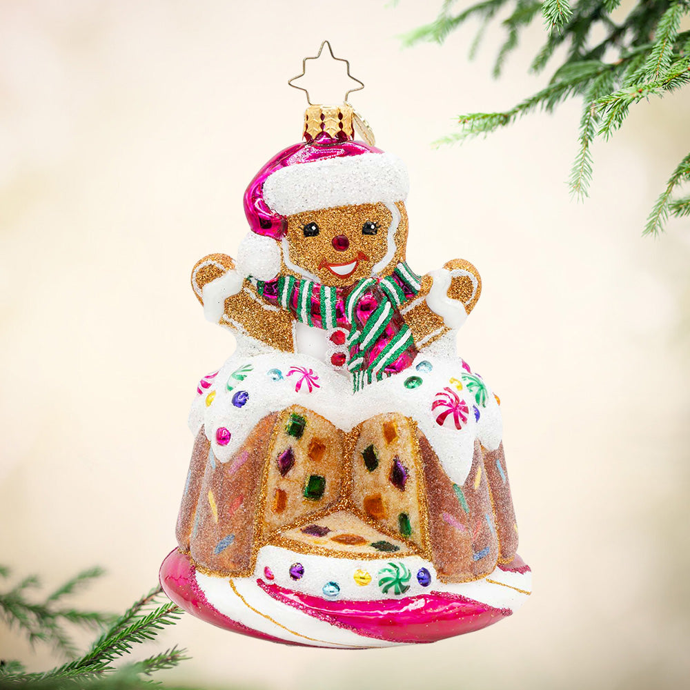 Bundt Surprise. Ornament of a gingerbread man in a pink hat and scarf popping out of a bundt cake with a slice missing and decorated in white frosting and colorful gems. View of front
