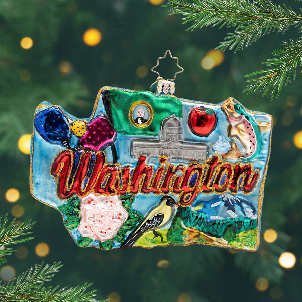 Washington Wonders. Ornament in the Shape of Washington state with the phrase "Washington" in red and gold, the states flag, a salmon, mountain, pink flowers, and the capital building with a blue background. View of front