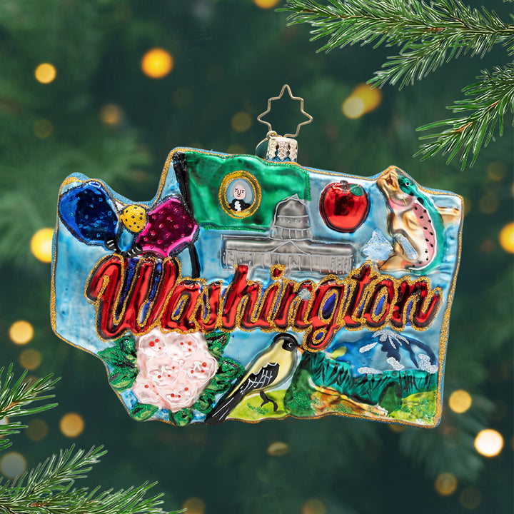 Washington Wonders. Ornament in the Shape of Washington state with the phrase "Washington" in red and gold, the states flag, a salmon, mountain, pink flowers, and the capital building with a blue background. View of front