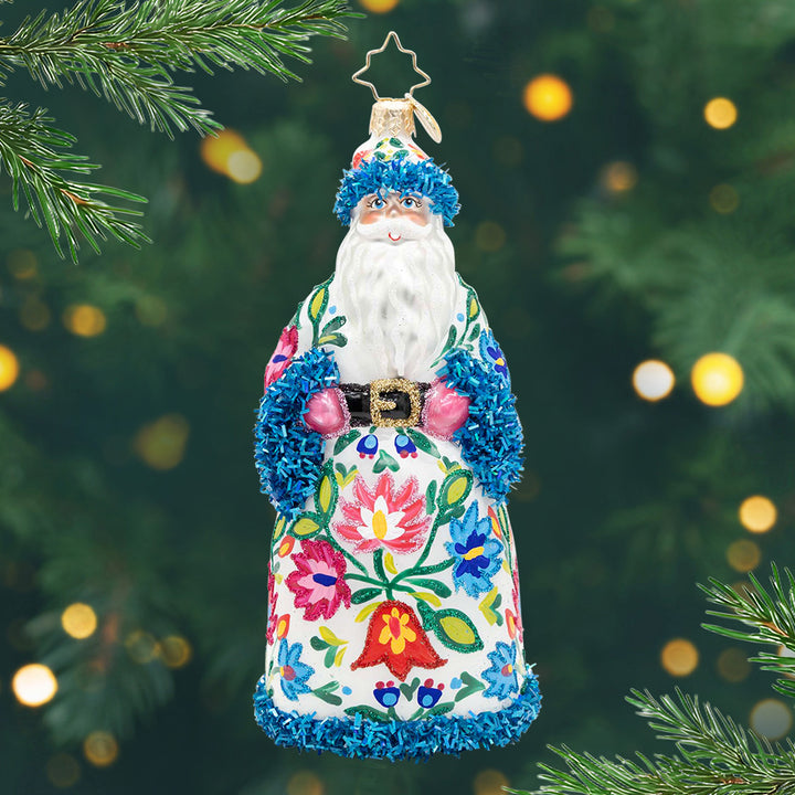 Folklore Noel. Santa ornament of him dressed in a white robe with blue sparkly trim and floral patterns. View of front