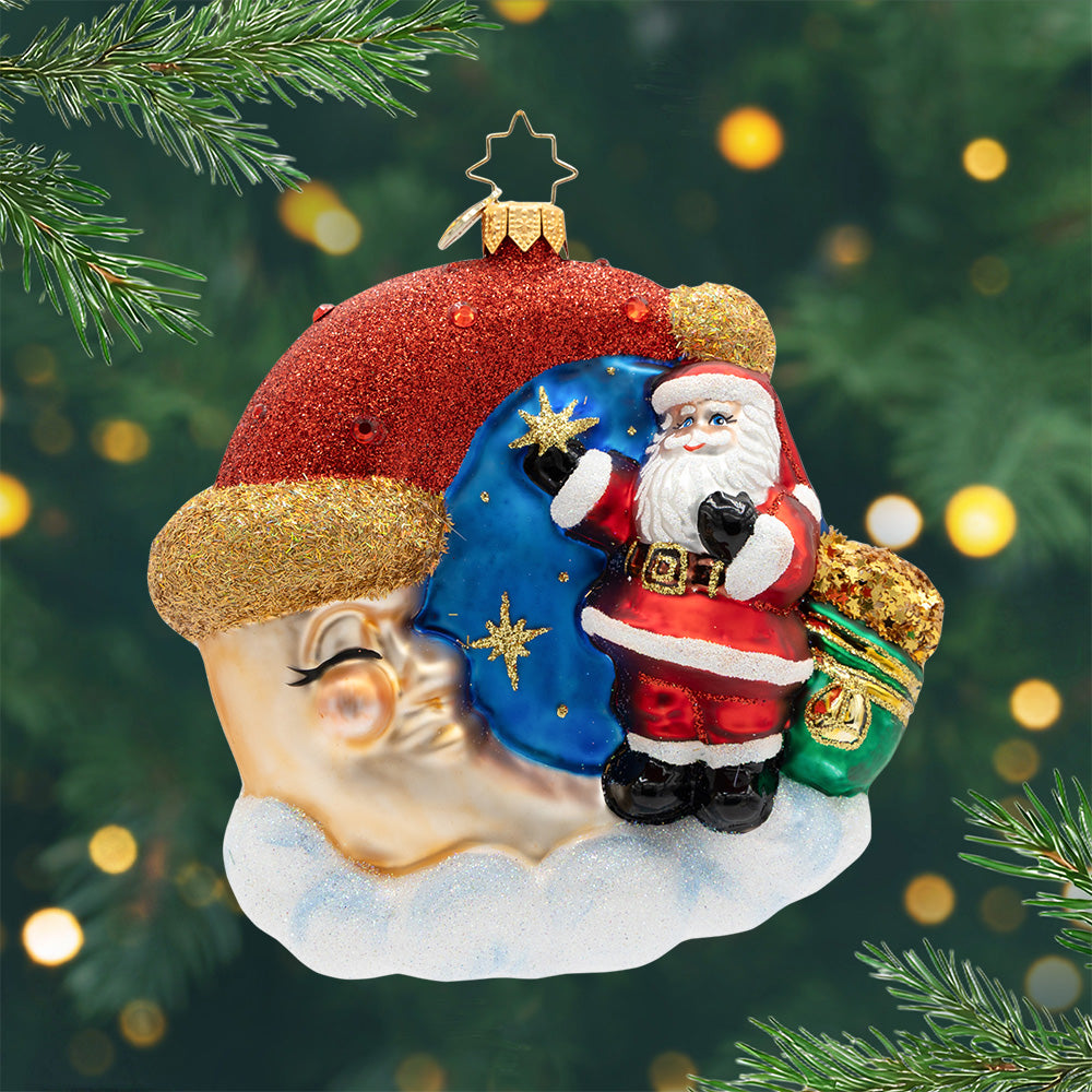 Crescent Wishes Santa. Ornament of Santa in his classic red suit and holing a green and gold bag outside next to a golden moon wearing a red Santa hat. View of front