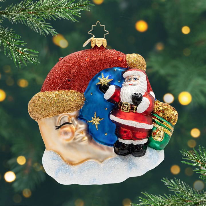 Crescent Wishes Santa. Ornament of Santa in his classic red suit and holing a green and gold bag outside next to a golden moon wearing a red Santa hat. View of front