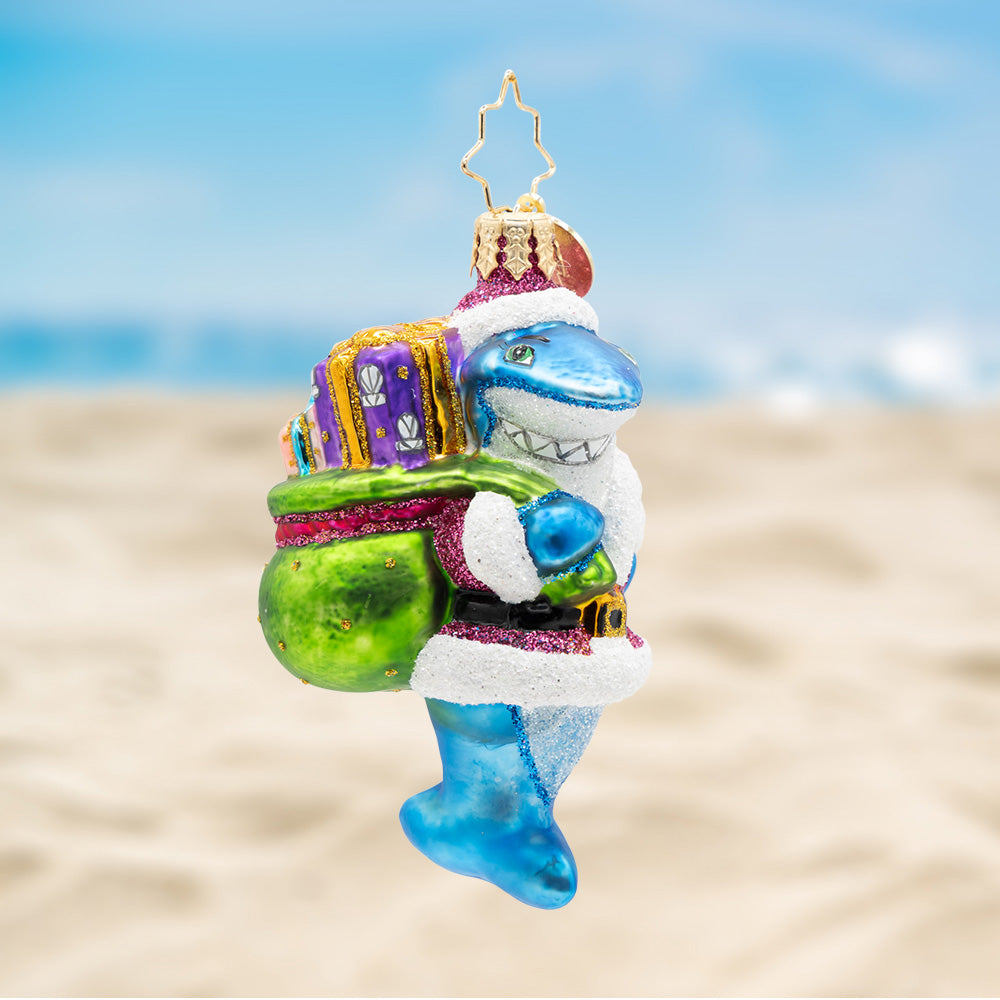 Santa Jaws Gem. Blue shark ornament dressed in a pink Santa suit, carrying a green bag with a purple present. View of front