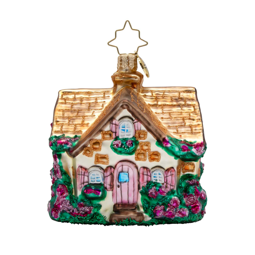 Floral Cottage Charm Gem. Ornament of a white cottage with a golden roof, a pink door and shutters, and dark green bushes with pink flowers. View of front.