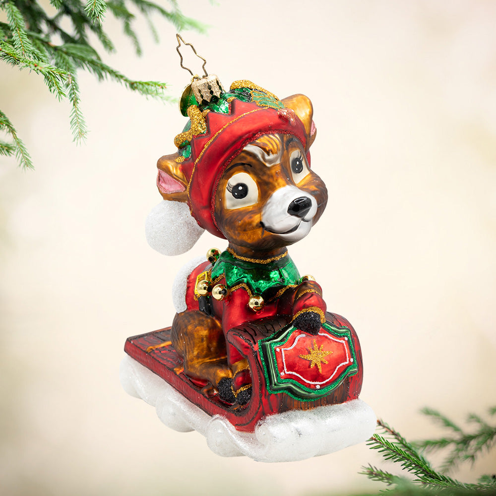 Sledding into the Season. Ornament of a reindeer dressed in a red and green elf suit on brown and red sled. View of front,