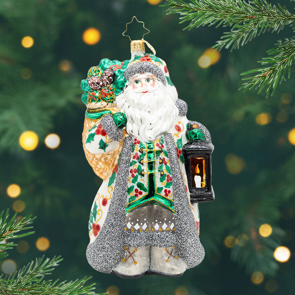Nordic Noel. Ornament of Santa in a white robe with mistletoes and gay sparkling trim while carrying a lantern and a bag with pinecones. View of front