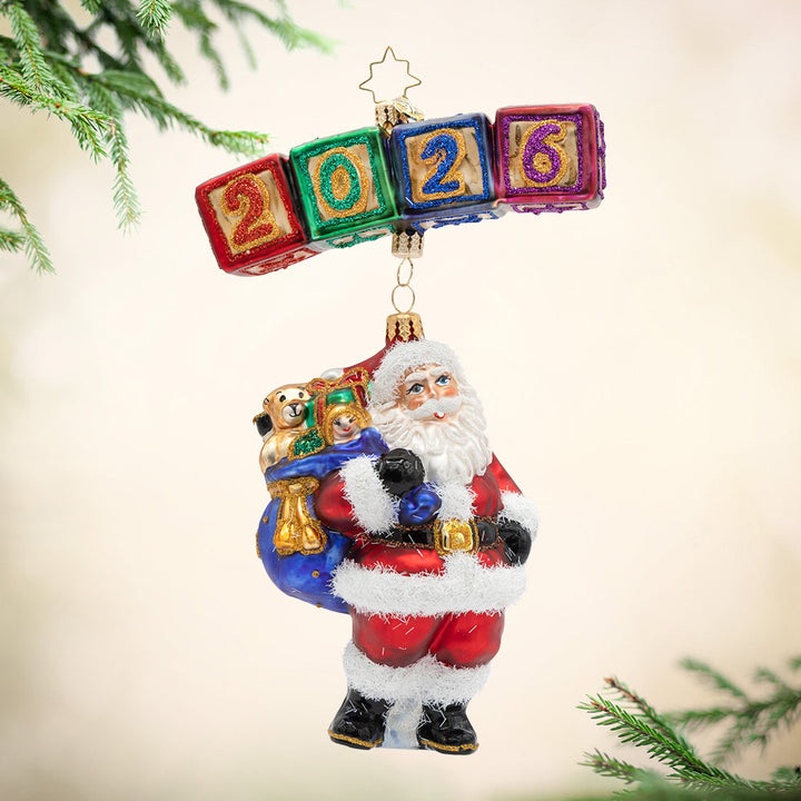 2026: Season of Smiles. Ornament of four colored blocks spelling 2026, holding Santa up with a blue bag of presents. View of front