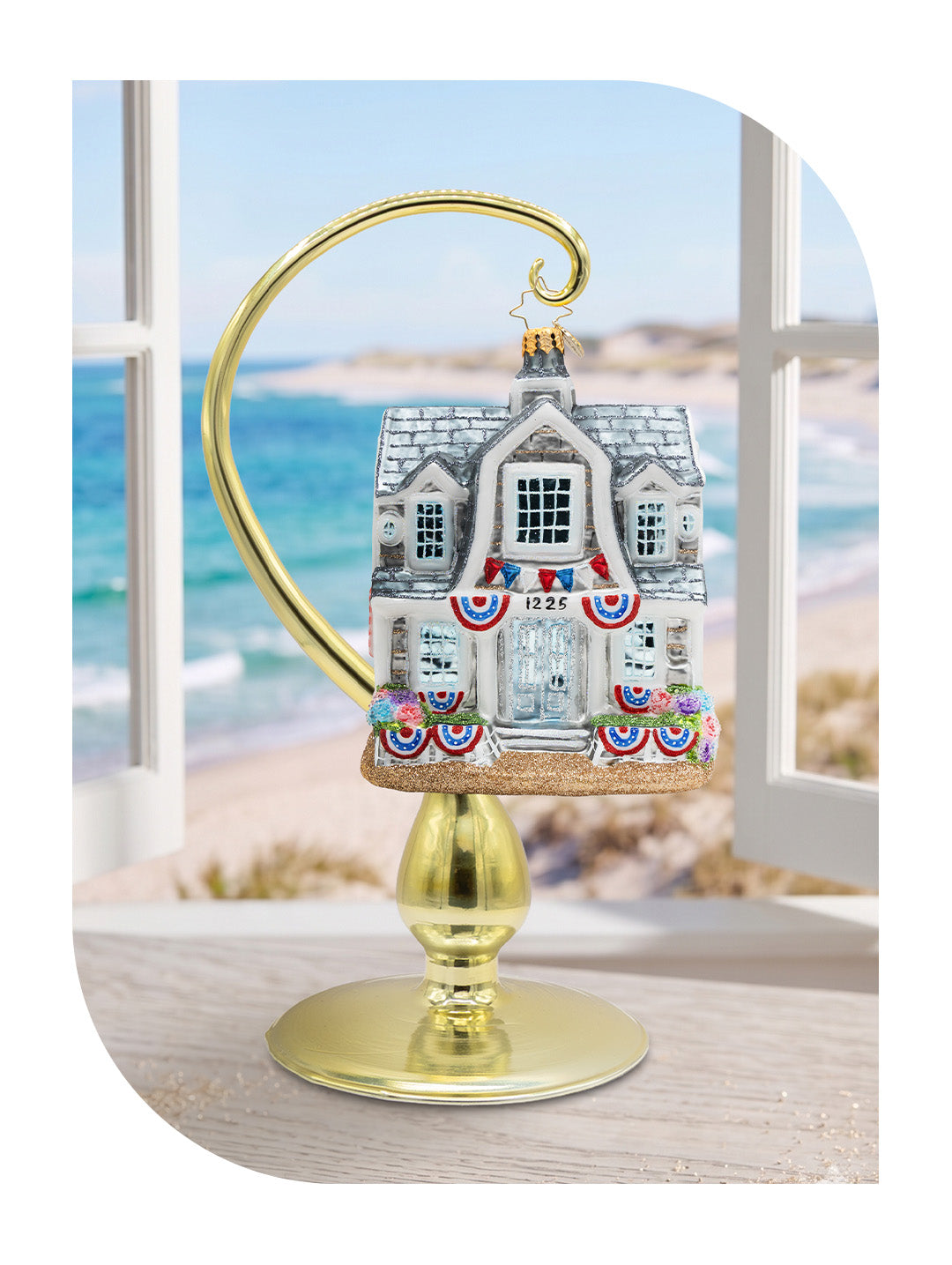 A glass ornament shaped like a beach house decorated with patriotic red, white, and blue bunting, displayed on a gold curved stand in front of a window overlooking a sandy beach.