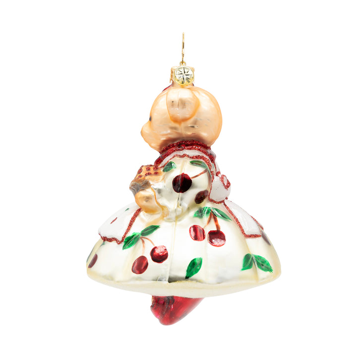 Side view of a bear ornament, dressed up in a white dress with cherries. Showcases a pie in the bear's arm. 