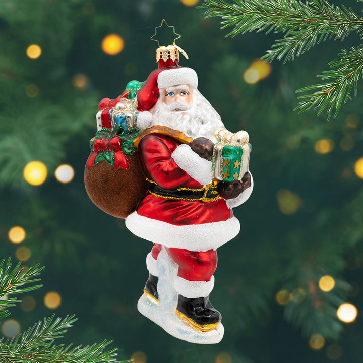 Merry Saint Nick. A Santa ornament of him in his classic red suit holding a brown bag of vibrant presents in one hand, and in the other a green present. View of front.