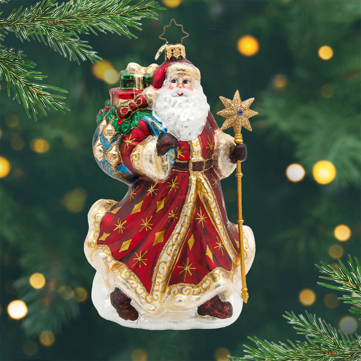 Starlight Keepsake Santa. Ornament of Santa in a red robe with gold accents is carrying a blue and white patterned bag full of presents while holding a gold staff. View of front.