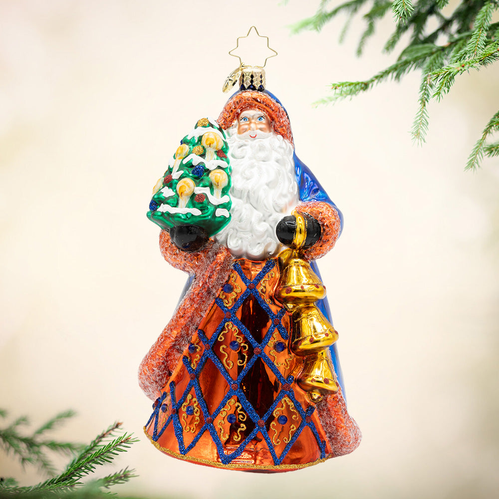 Royal Tidings Santa. This ornament of Santa dressed in a magnificent blue and copper coat, he holds a decorated Christmas tree and golden bells. View of front.