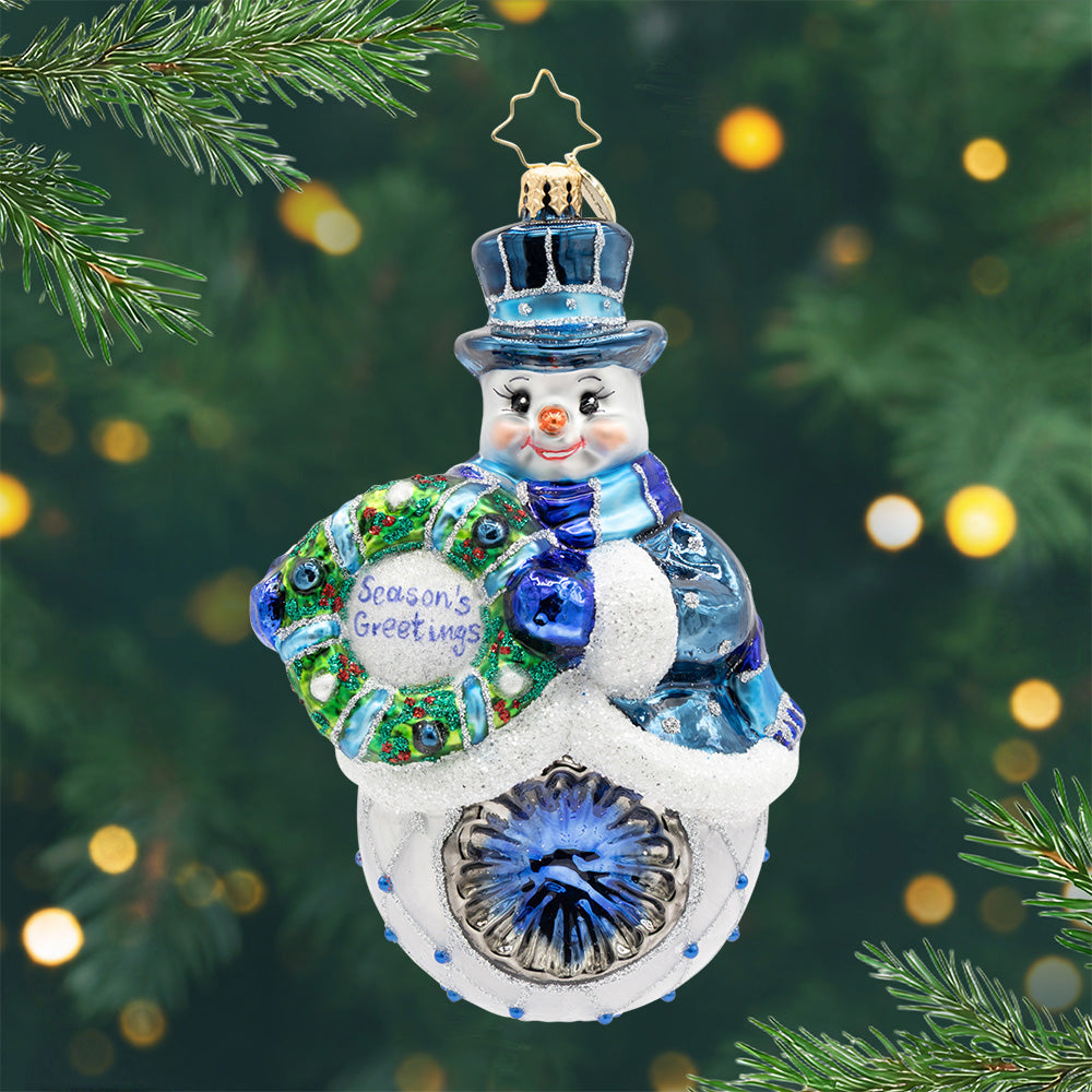 Wreathfully Yours. A snowman ornament dressed up in a top hat, scarf, and coat, all in shades and gems of blue and silver while holding a green wreath with the phrase of seasons greetings. View of front.