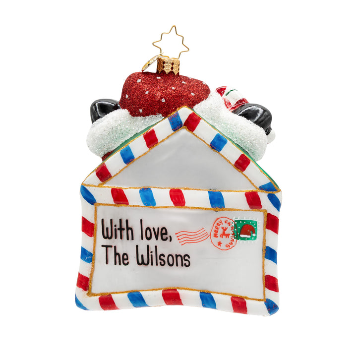 A personalized Christmas ornament shaped like a house envelope, decorated with red and blue striped borders, a red glittery hat on top, and the text 'With love, The Wilsons' printed on the front.