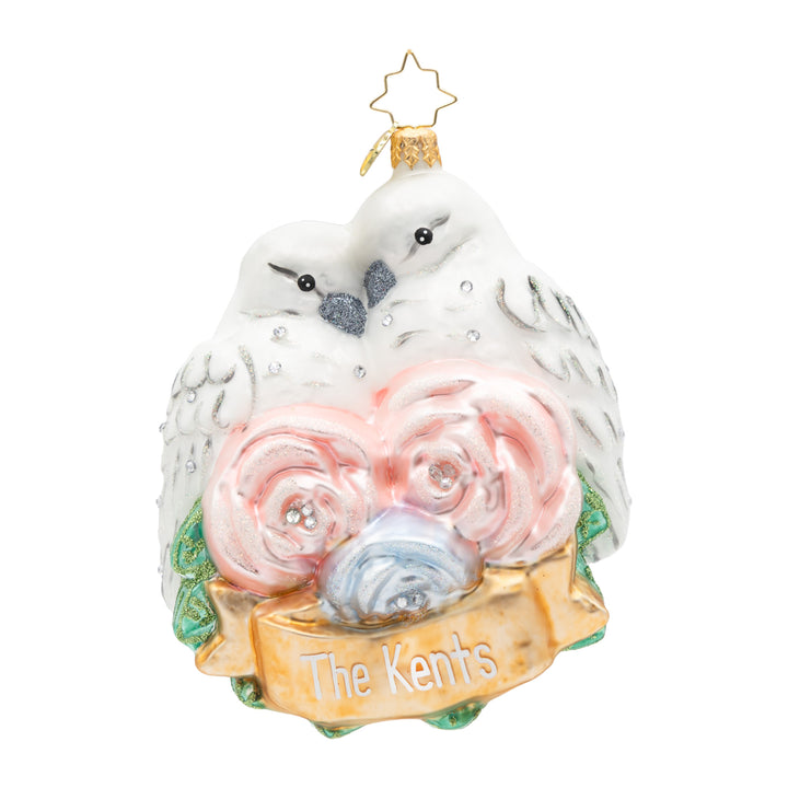 A personalized Christmas ornament shaped like two white doves nesting among pink and blue roses, with a gold banner reading 'The Kents' and a gold hanging loop at the top.