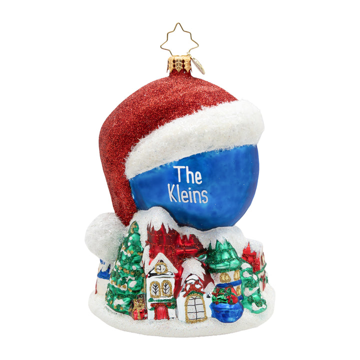 A personalized Christmas ornament shaped like a Santa hat with a snowy white brim, a glittery red top, and a blue center printed with the name 'The Kleins' in white. The bottom features a small multicolor winter village scene with houses, trees, and gifts on a white base, topped with a gold star loop.