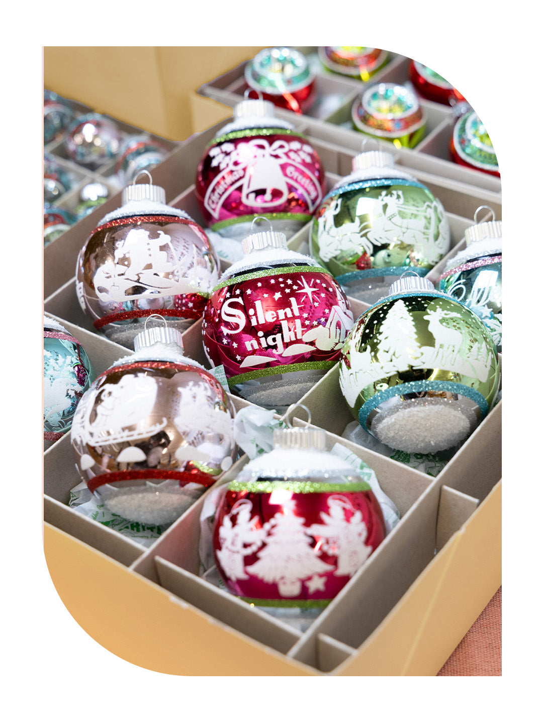 An assortment of glass Christmas ornaments in a display box, featuring holiday motifs like bells, snowmen, and reindeer.
