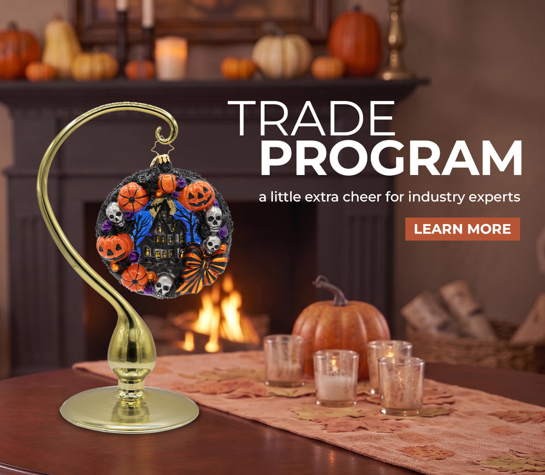 A Halloween-themed glass ornament featuring pumpkins, ghosts, and a haunted house design, displayed on a gold stand near a fireplace with pumpkins and candles, promoting the Trade Program with a 'Learn More' button.