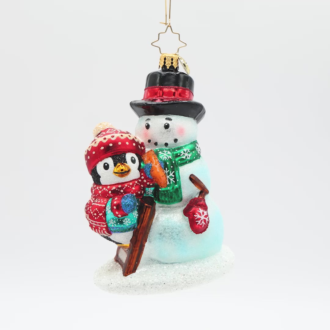 Front view of a penguin ornament wearing a blue coat and a matching red hat and scarf, finishing decorating the snowman it built.