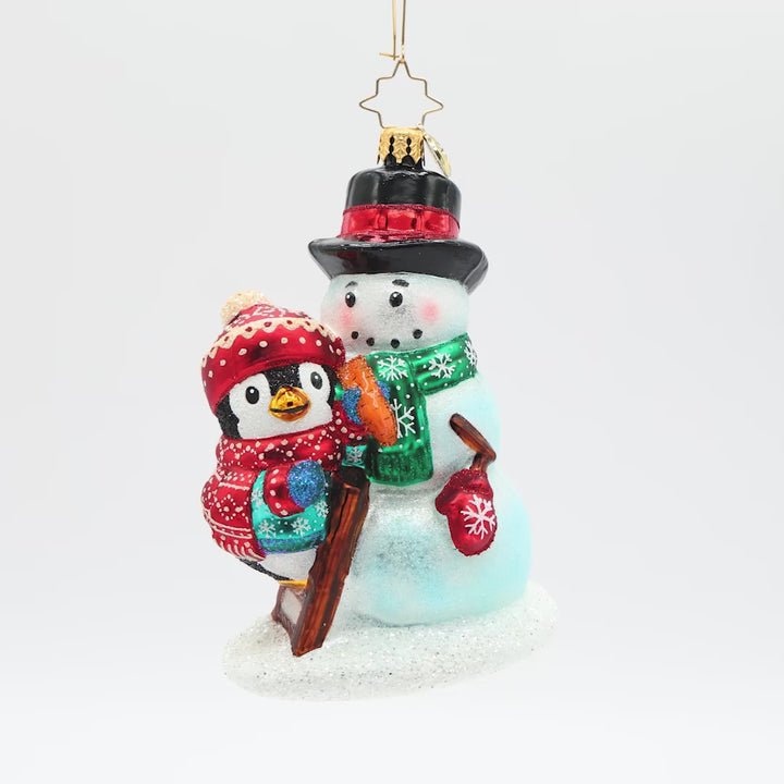 Front view of a penguin ornament wearing a blue coat and a matching red hat and scarf, finishing decorating the snowman it built.